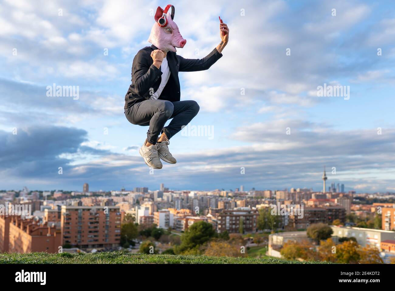 Homme professionnel portant un masque de porc prenant selfie de smartphone tout en sautant dans la ville contre le ciel Banque D'Images