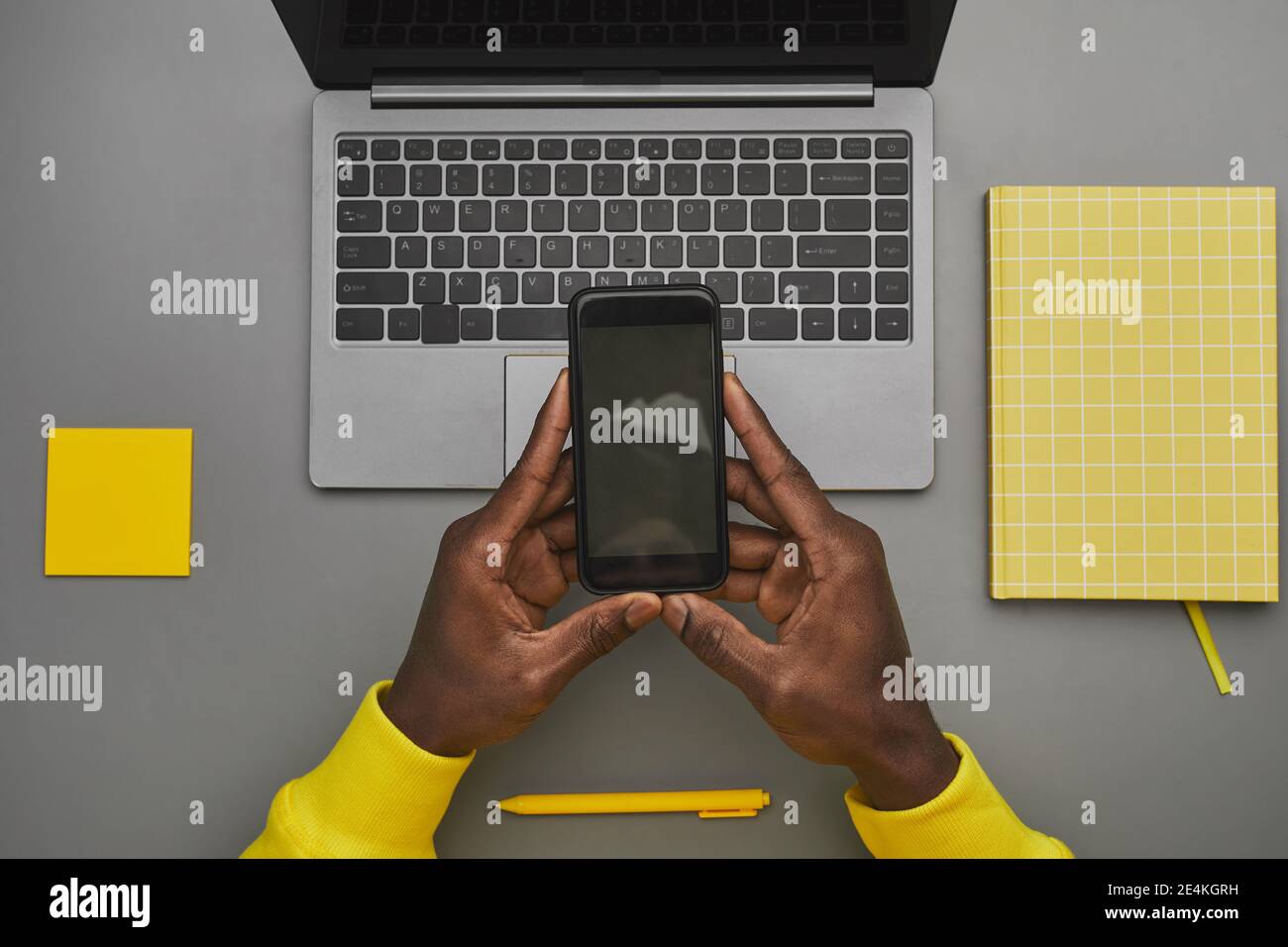 Arrière-plan gris et jaune représentant des mains afro-américaines de sexe masculin tenant un smartphone avec un écran vide sur un ordinateur portable tout en travaillant à un bureau, de haut en bas vie Banque D'Images