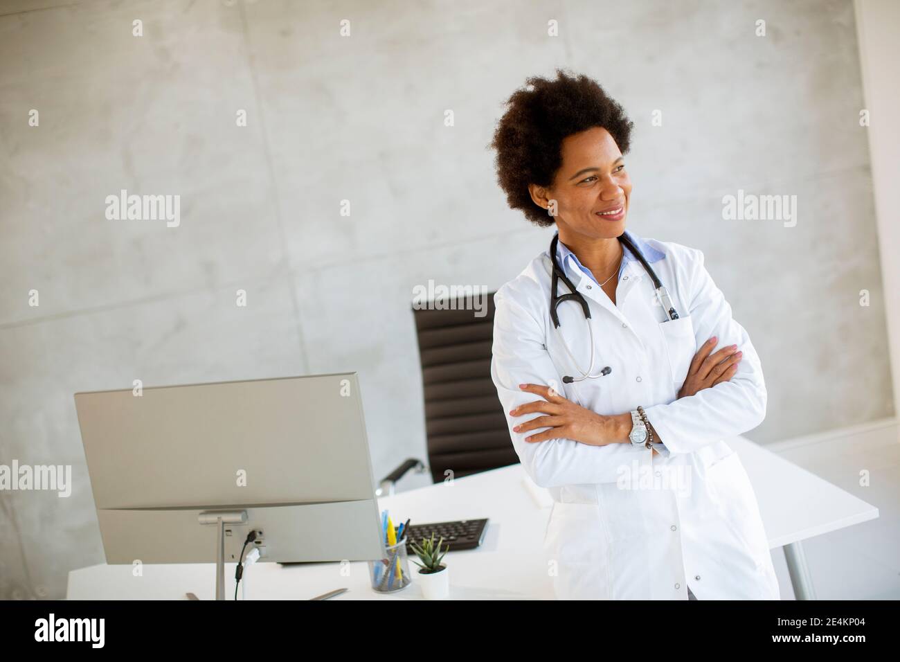Femme afro-américaine médecin portant un manteau blanc avec stéthoscope debout par bureau dans le bureau Banque D'Images