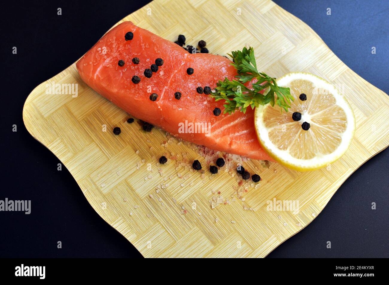 Une tranche de saumon cru assaisonnée de persil, de poivre, de sel et de citron sur un hachoir en bois et un fond noir. Vue de dessus Banque D'Images