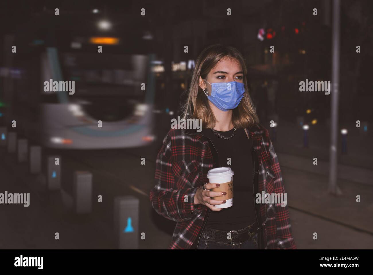 Une adolescente avec un masque facial médical marchant dans une rue avec un café à emporter sur sa main, il y a un tram en arrière-plan la nuit. Banque D'Images