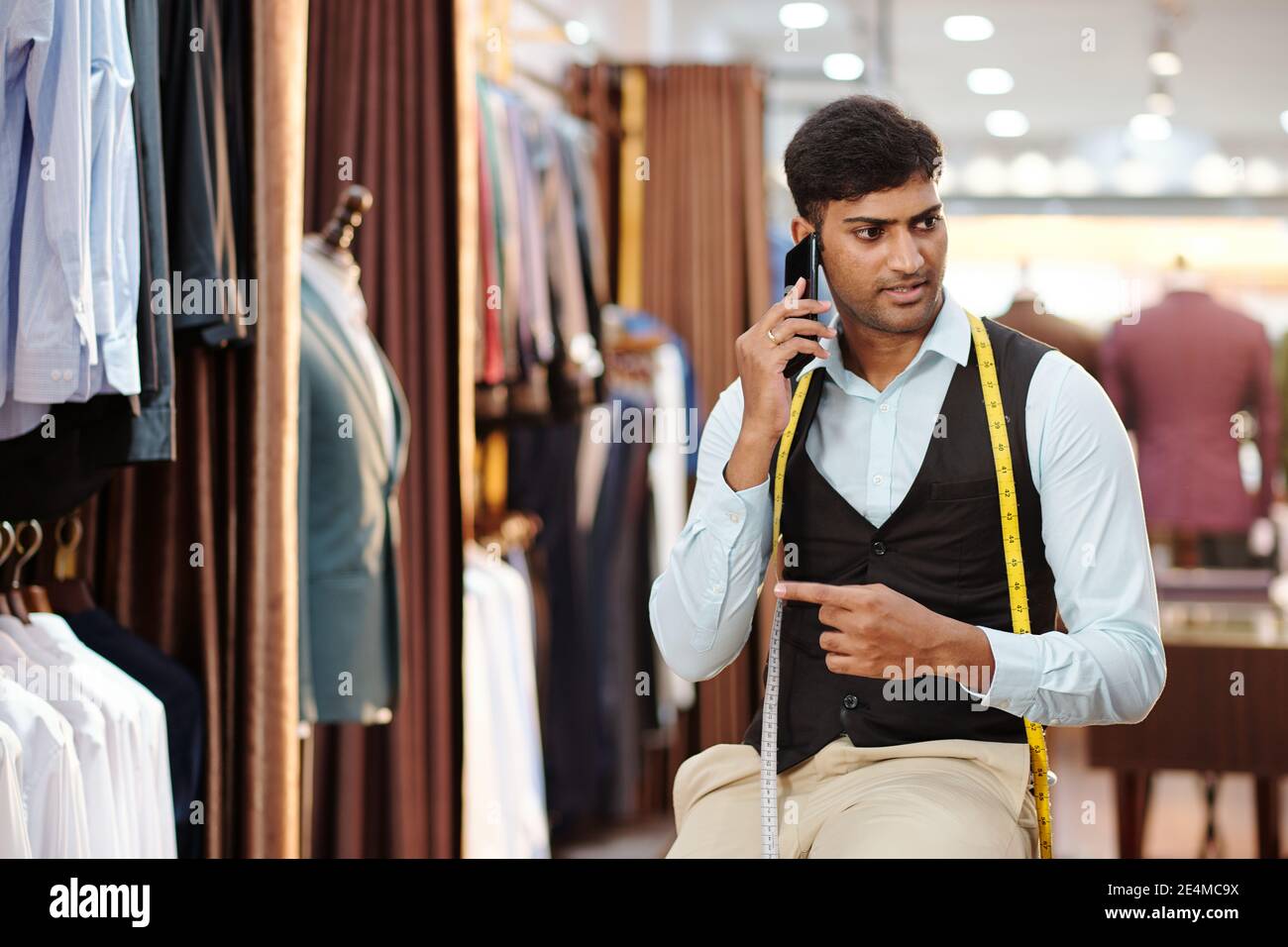 Portrait d'un jeune Indien sérieux tailleur travaillant dans l'atelier de mode et parler au téléphone avec le client Banque D'Images