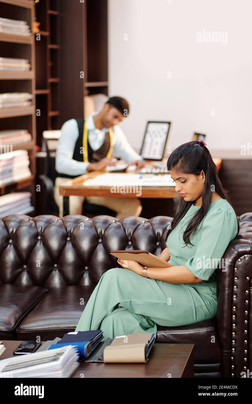 Jeune directeur d'atelier indien assis sur un canapé et lisant des articles et les e-mails des clients sur une tablette lors du calcul sur mesure consommation de tissu Banque D'Images