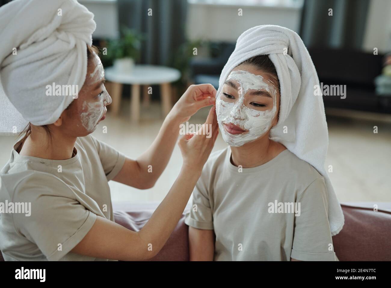 Jeunes femmes jumeaux avec masque en argile facial assis sur le canapé dans l'environnement à la maison tandis que l'un d'entre eux aide sa sœur placez une serviette sur la tête Banque D'Images