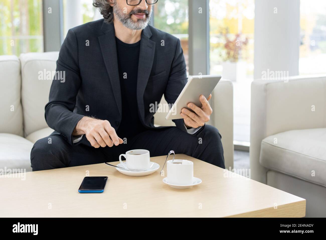 Courtier à barbe mature en tenue de coffee ayant du café et regardant à travers nouvelles en ligne tout en passant du temps dans un café d'affaires modernes centre Banque D'Images