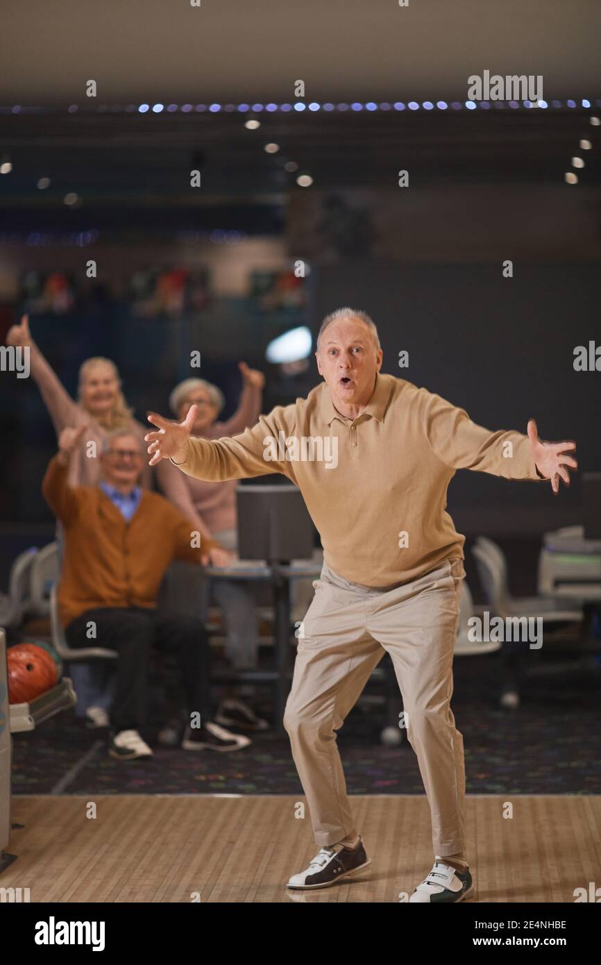 Portrait vertical complet d'un homme senior enthousiaste jouant au bowling et célébrer la victoire ou la grève avec un groupe d'amis dans arrière-plan Banque D'Images