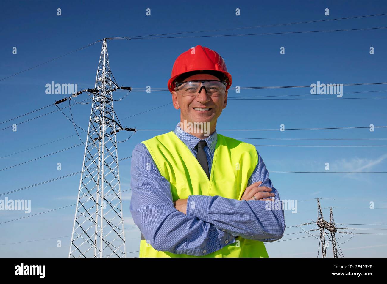Ingénieur souriant dans Red HardHat et Yellow West debout sous les tours de transmission électrique. Happy Manager dans Red HardHat et Yellow Vest. Banque D'Images