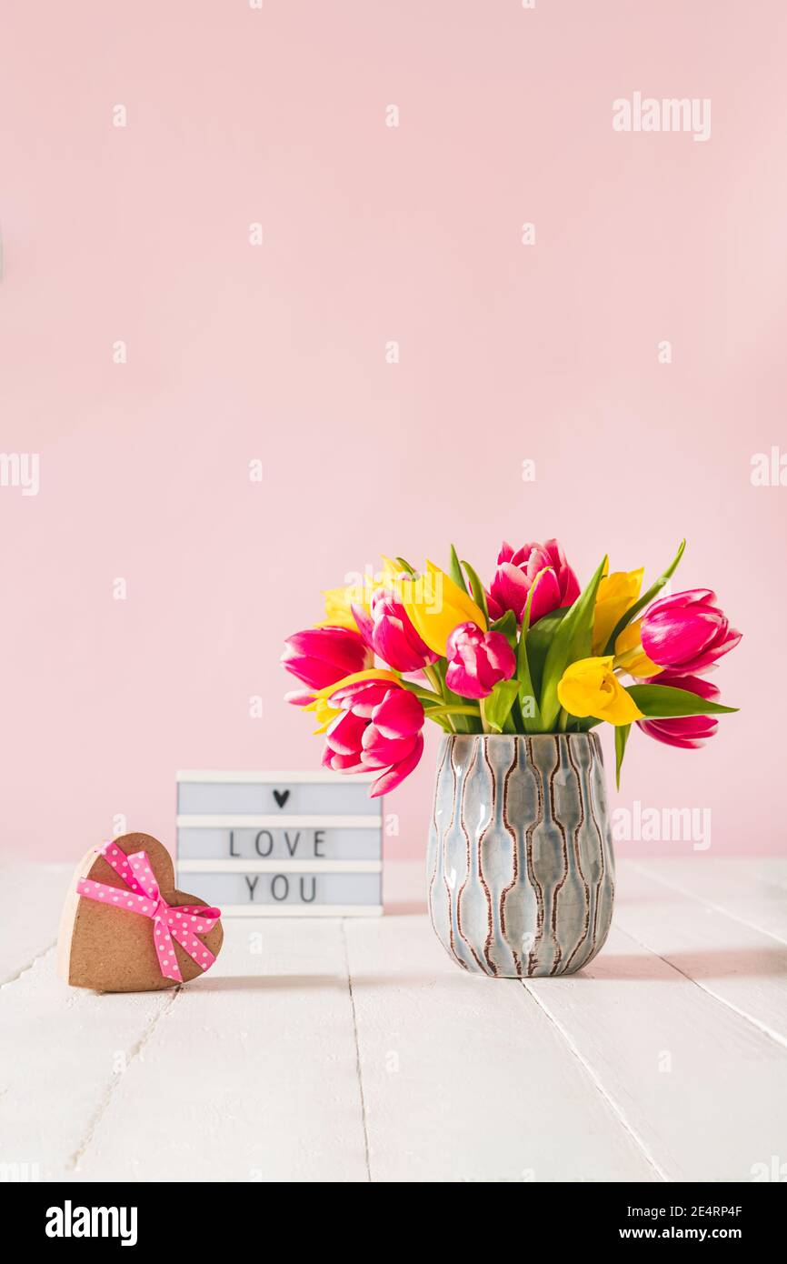 Boîte cadeau en forme de coeur recyclé, bouquet de printemps de fleurs de tulipe roses et jaunes dans un vase et une visionneuse avec message d'amour sur la table blanche en bois Banque D'Images