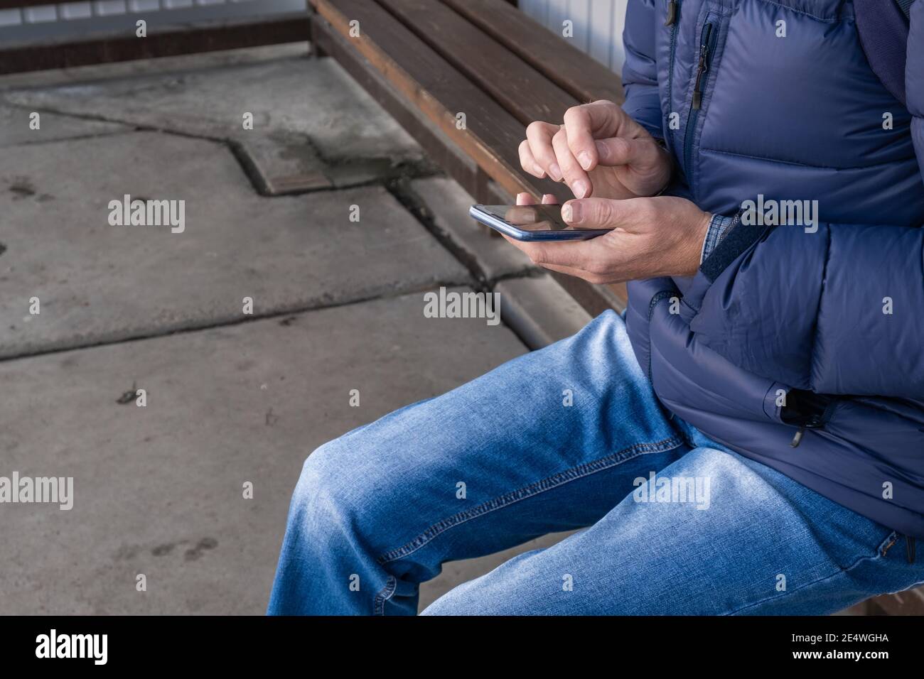 Homme en veste et jeans est assis à l'arrêt des transports en commun et attend le bus.appelle le taxi.en utilisant son smartphone, il suit les informations sur l'emplacement Banque D'Images