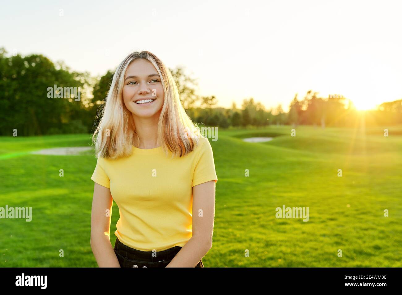 Portrait extérieur d'une adolescente souriante de 16 ans, 17 ans en T-shirt jaune, sur une pelouse verte ensoleillée Banque D'Images