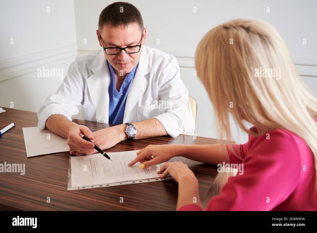 Chirurgien masculin parlant avec une patiente au bureau. Jeune femme blonde discutant de chirurgie plastique avec un médecin dans une clinique moderne. Concept de rendez-vous médical et de chirurgie esthétique. Banque D'Images