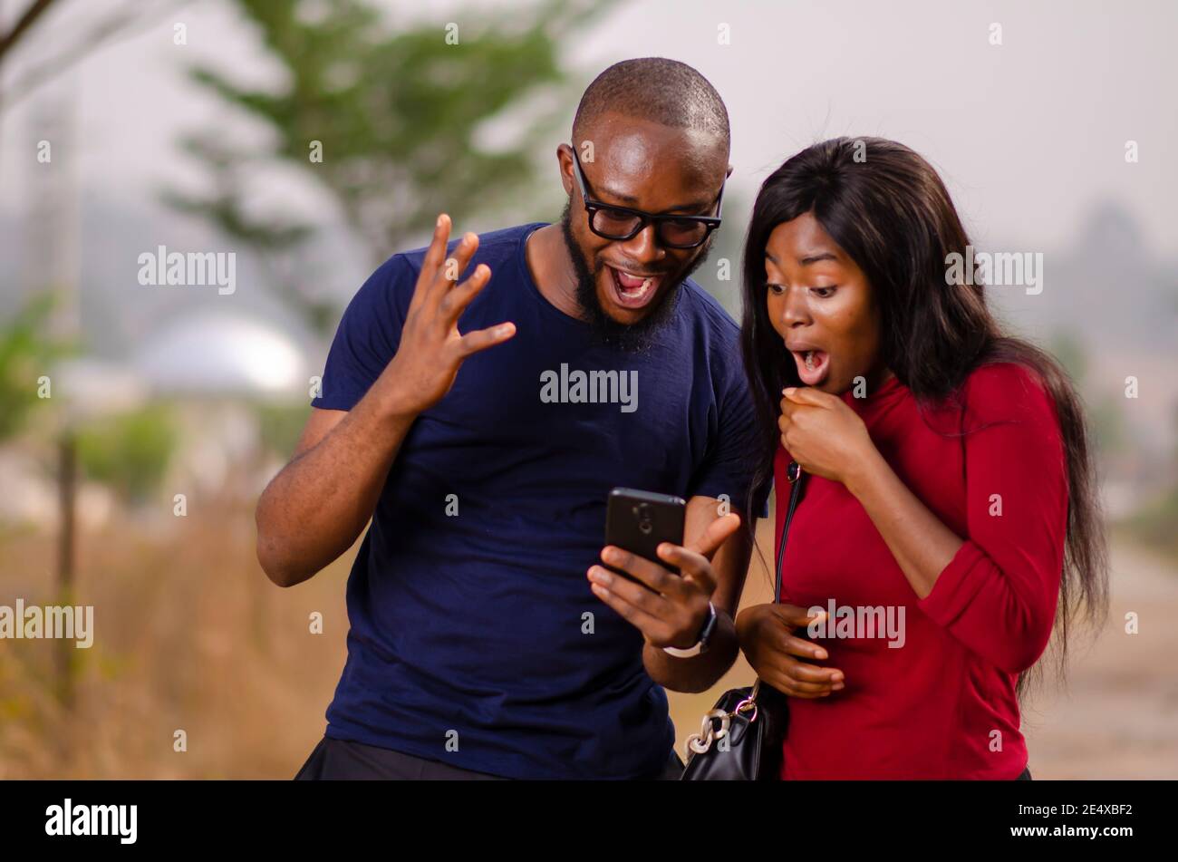 jeune homme et femme africains debout dehors et enchantés pendant à l'aide de leur smartphone Banque D'Images