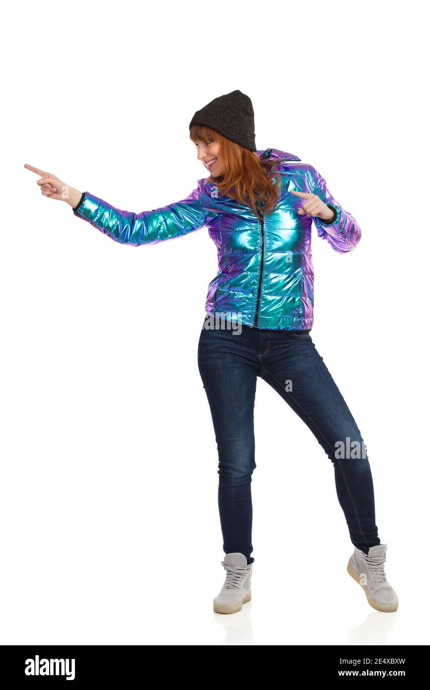 Bonne jeune femme en duvet, casquette, jeans et baskets brillants et éclatants, regarde loin et touche quelque chose avec l'index. Goujon sur toute la longueur Banque D'Images