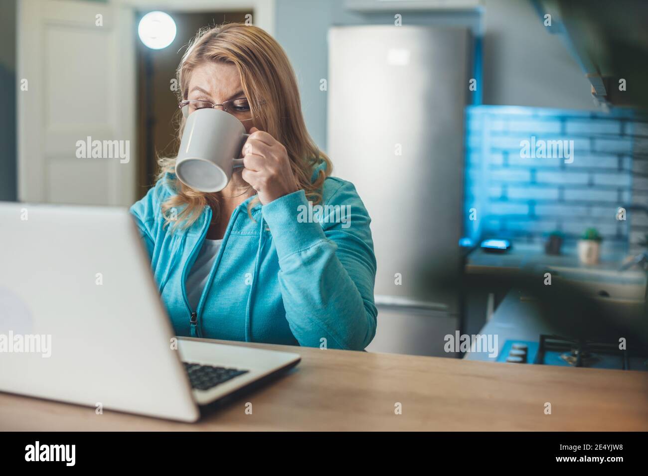 Une femme blonde âgée occupée boit une tasse de thé tout en travaillant à la maison à l'ordinateur portable dans la cuisine Banque D'Images