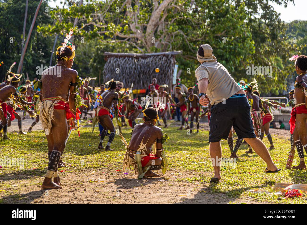 Un passager du navire d'expédition True North lance la balle orange sur le gardien de wicket accroupi lors d'une partie de cricket de style des îles Trobriand à Kwebwaga, îles Trobriand, Papouasie-Nouvelle-Guinée, un danseur costumé se tenant à ses côtés, d'autres danseurs à l'arrière-plan devant une hutte en chaume de palmiers. Banque D'Images