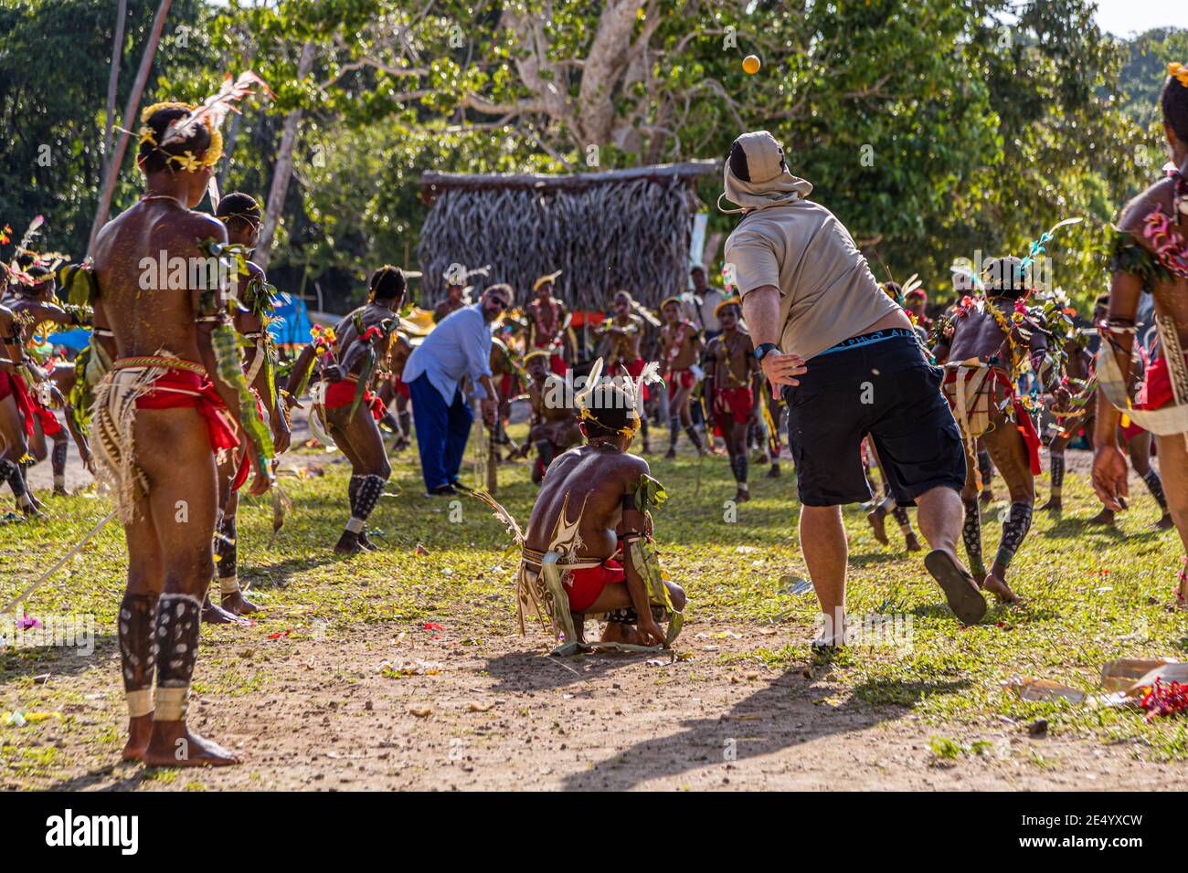 Un passager du navire d'expédition True North, vêtu d'une chemise blanche et d'un pantalon bleu, lance la balle orange lors d'une partie de cricket dans le style des îles Trobriand à Kwebwaga, îles Trobriand, Papouasie-Nouvelle-Guinée, un danseur en costume accroupi devant le guichet, des hommes en costume dansant autour d'eux. Banque D'Images