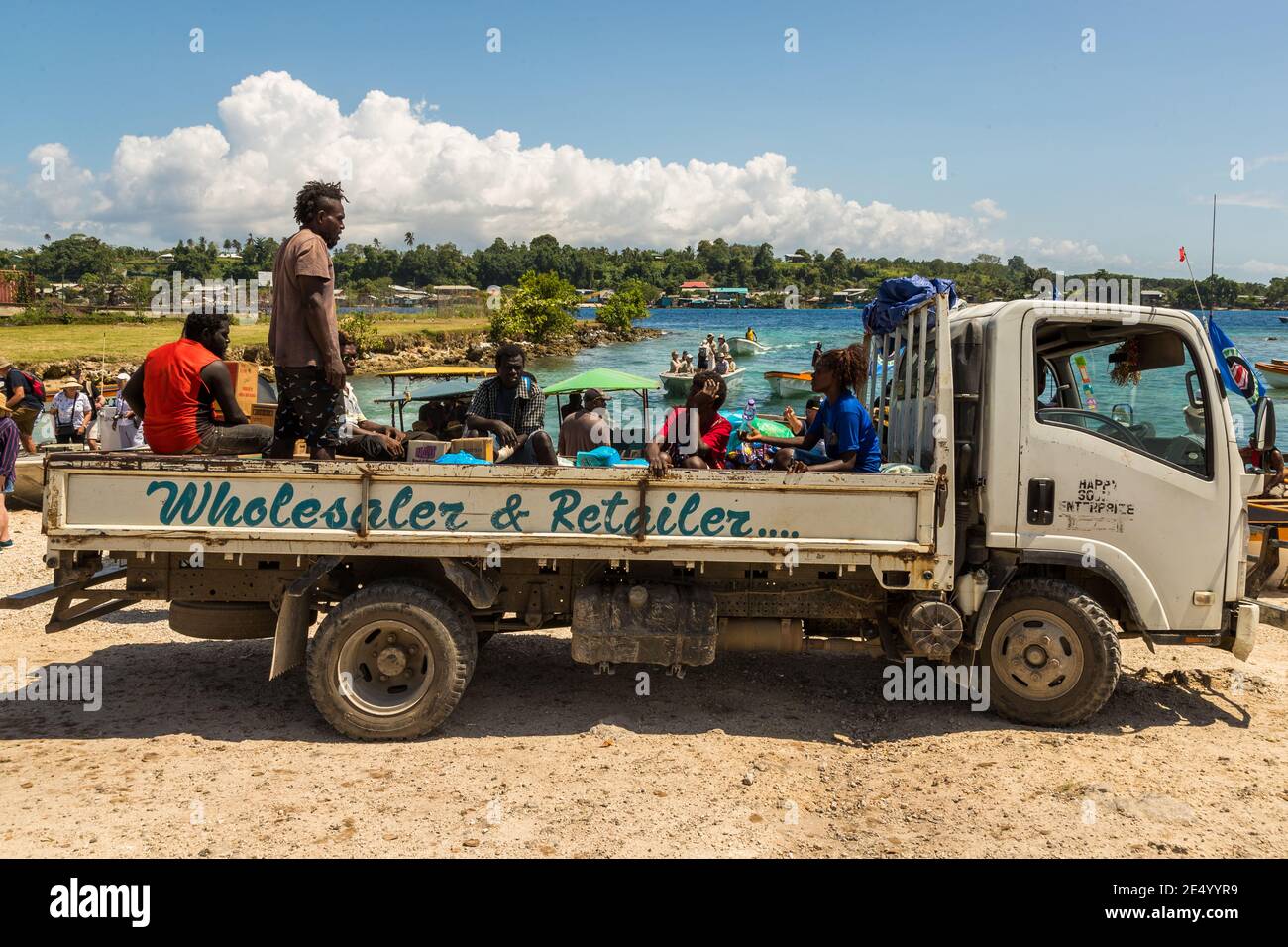 Un camion portant l'inscription "Grossiste & Détail" transporte de nombreux passagers sur sa plate-forme de chargement à Buka, Bougainville, Papouasie-Nouvelle-Guinée. Des bateaux sont amarrés en arrière-plan, d'autres personnes sont assises sous un parasol vert. Banque D'Images