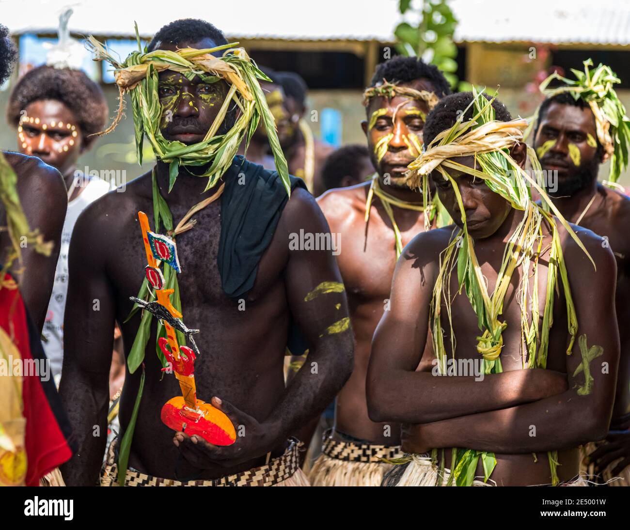 Un homme au visage peint en vert et portant une coiffe en feuilles de palmier tient un objet en bois plus petit, peint de la même manière, lors d'un festival du village Sing-Sing à Bougainville, Papouasie-Nouvelle-Guinée, d'autres hommes portant des ornements en herbe autour du cou en arrière-plan. Banque D'Images