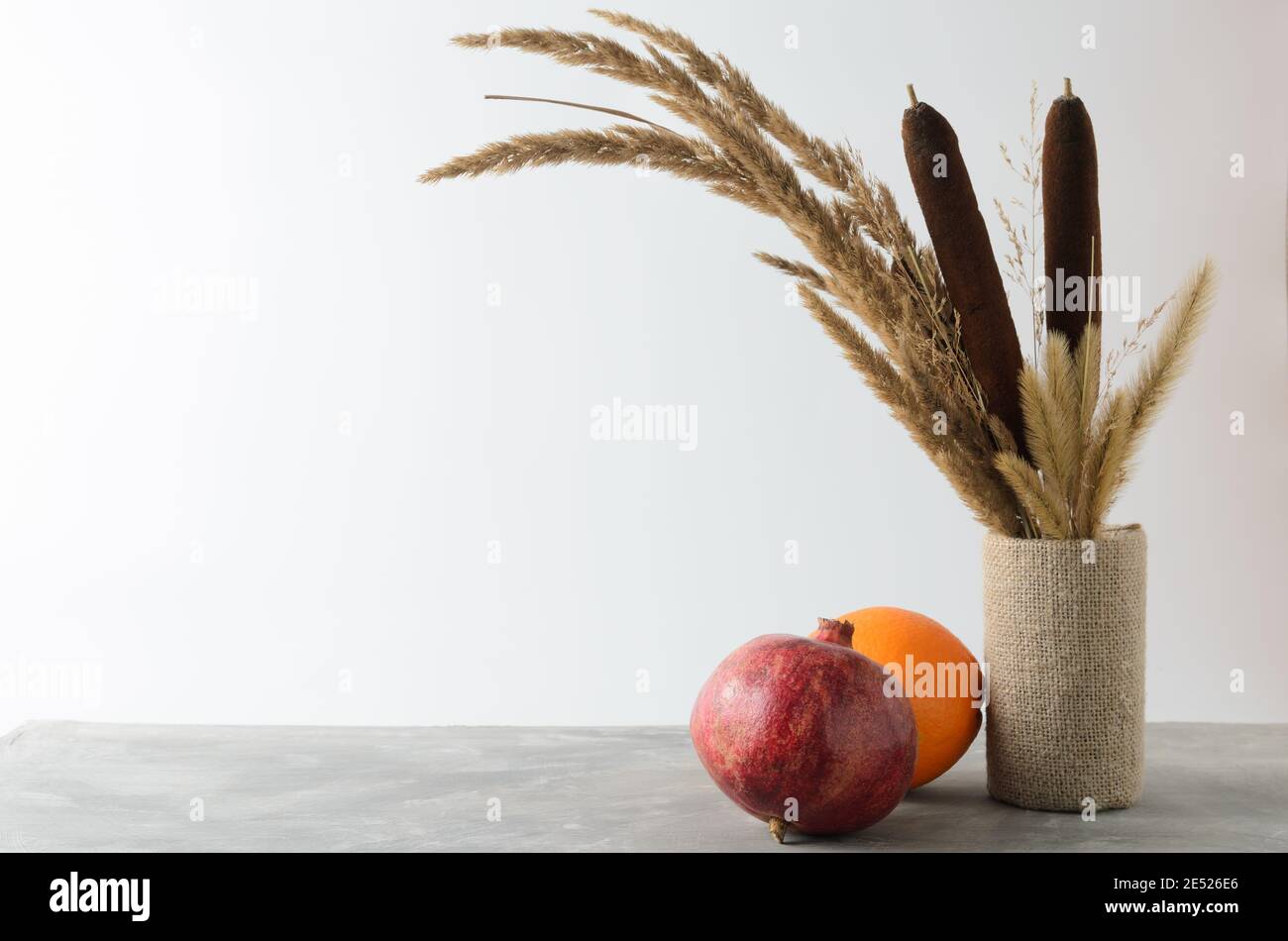 Une orange et une grenade sur une table grise texturée contre un mur blanc avec un bouquet d'automne d'herbes séchées et de roseaux. Mise au point sélective. Banque D'Images
