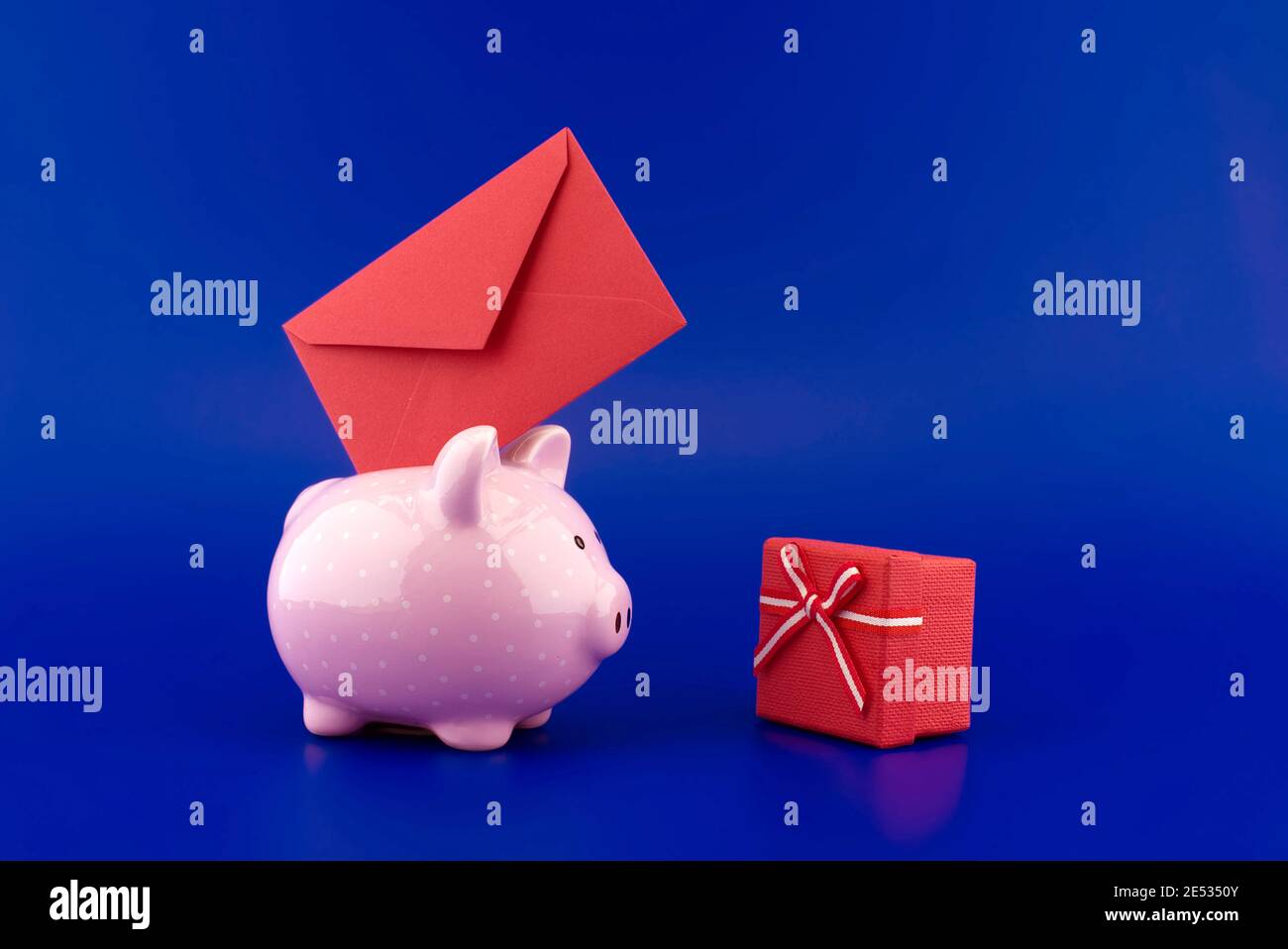Enveloppe rouge festive insérée dans une tirelire rose à côté d'une boîte de présentation sur fond bleu avec copie espace Banque D'Images