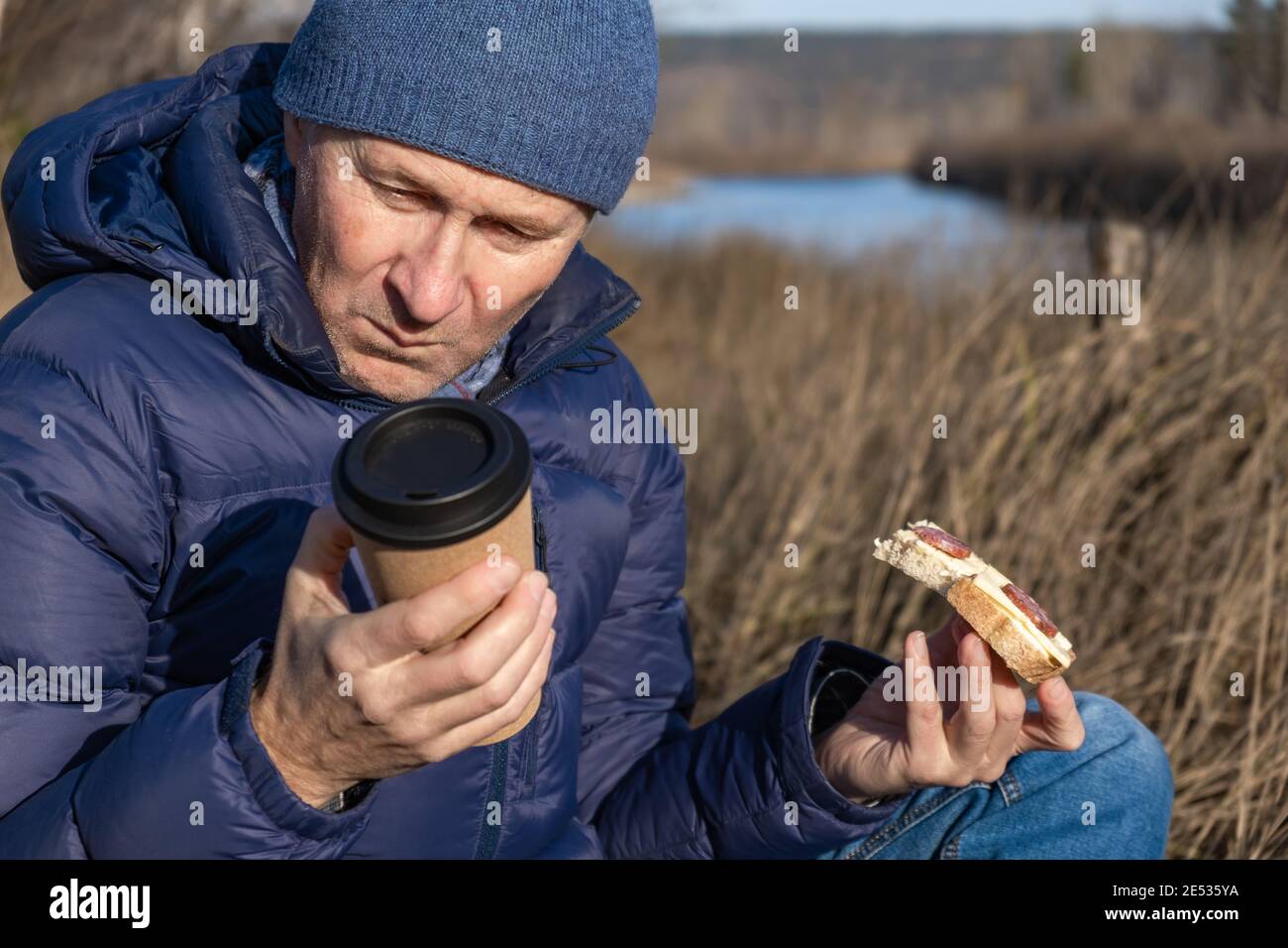 À l'automne, un homme s'assoit sur l'herbe, mange un sandwich avec du fromage et des saucisses et regarde un verre de café. Un en-cas rapide et savoureux en marchant. Banque D'Images