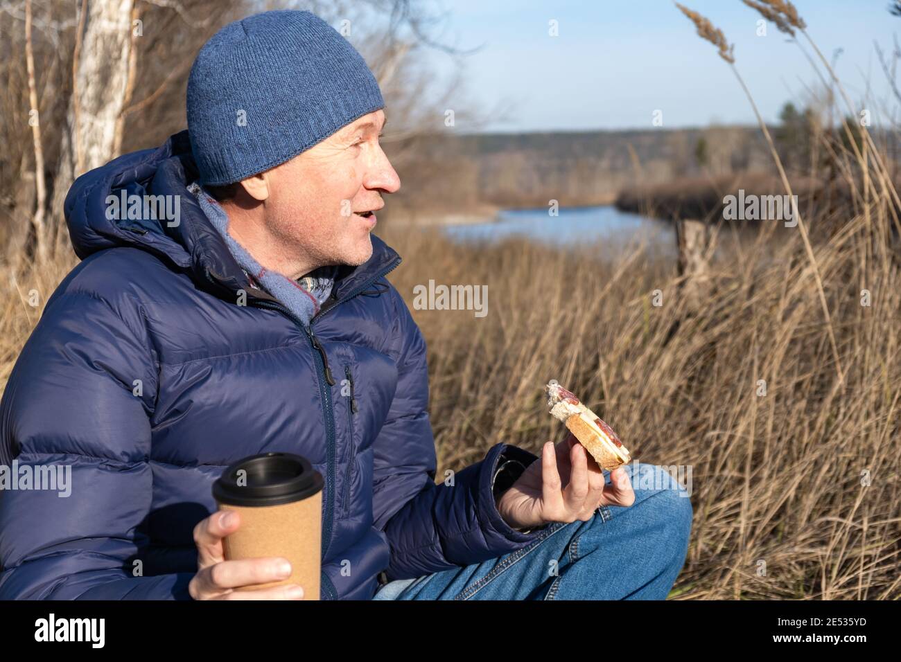 À l'automne, un homme s'assoit sur l'herbe, mange un sandwich avec du fromage et des saucisses, sourit et boit du café. Un en-cas rapide et savoureux en marchant. Banque D'Images