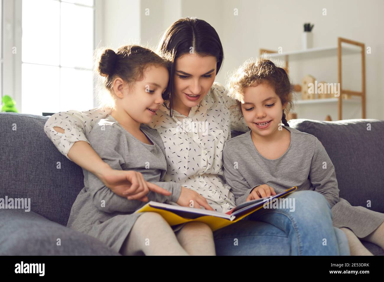 Bonne jeune mère lisant un livre assis à la maison sur le canapé avec ses filles jumelles. Banque D'Images