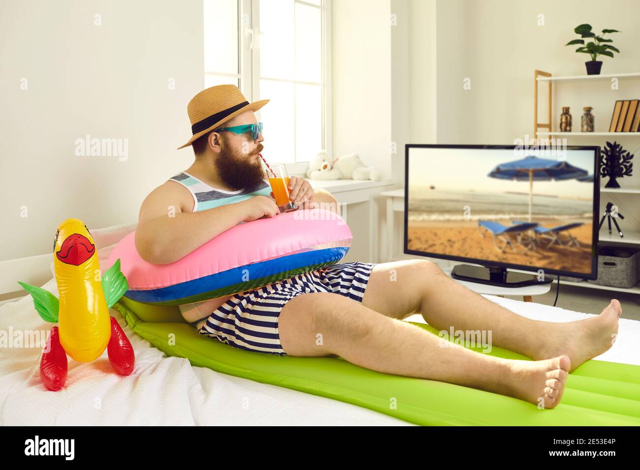 Drôle d'homme avec des jouets gonflables de plage sirotant un cocktail et regarder Émission de télévision de voyage en toute verrouillage Banque D'Images