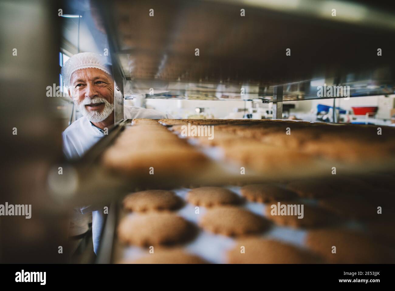 Photo d'homme boulanger adulte professionnel en uniforme de travail blanc. Debout devant les étagères avec des petits gâteaux frais. Banque D'Images