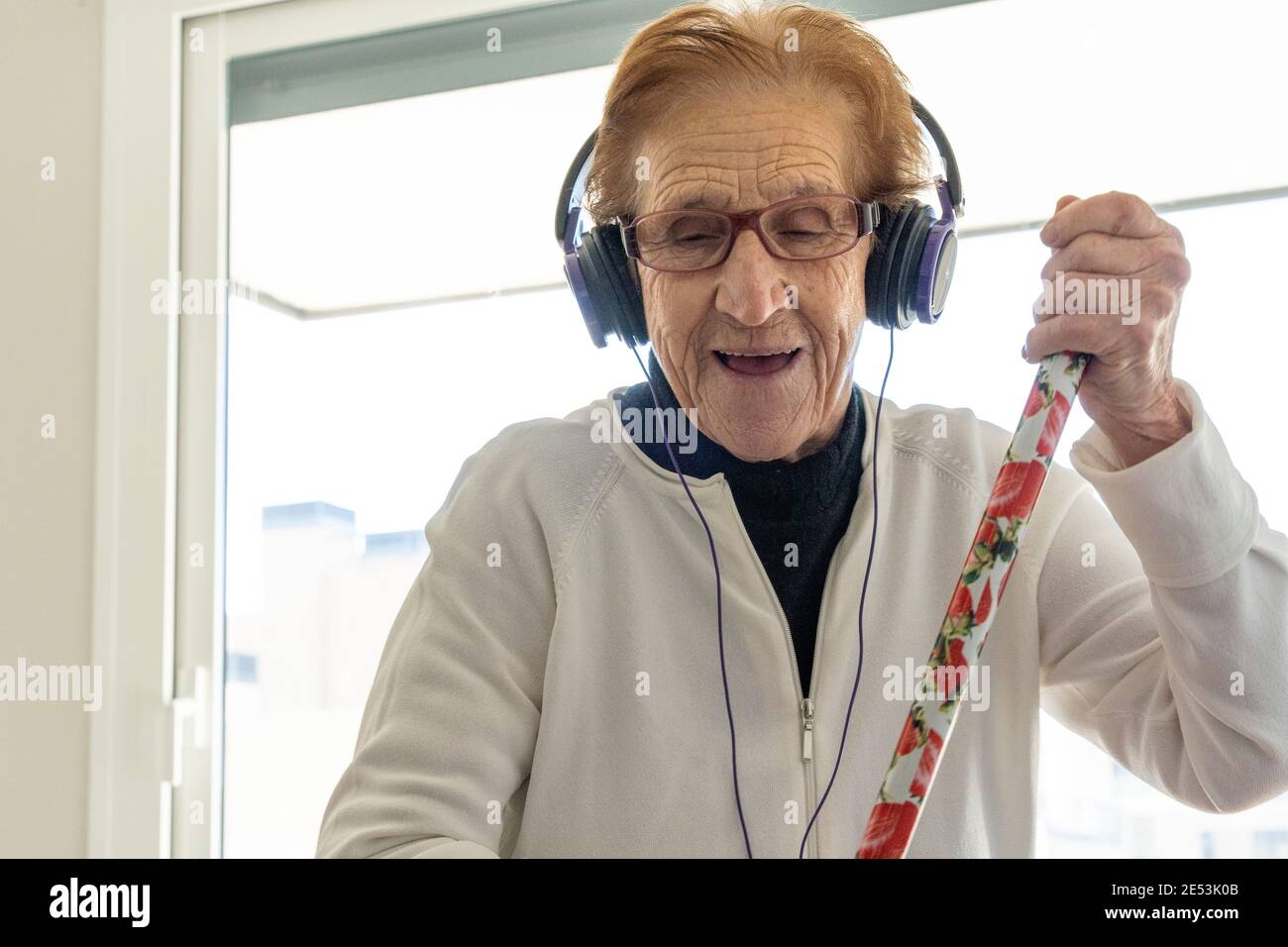 Femme âgée optimiste dans un casque d'écoute, écoutant de la musique et appréciant chansons tout en faisant des travaux ménagers avec un balai Banque D'Images