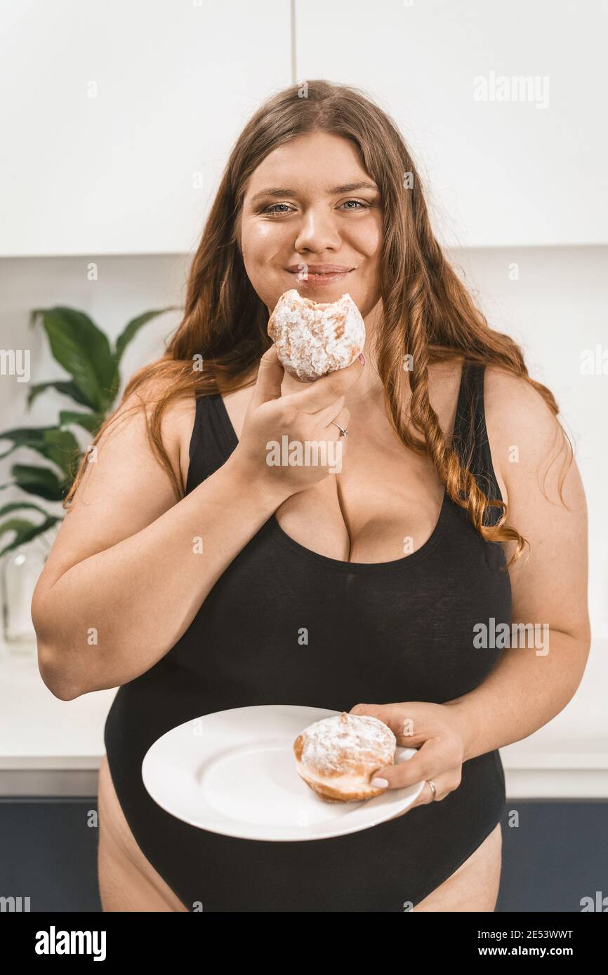 Satisfait jeune femme gras mangeant un gâteau souriant heureux sur l'appareil photo portant un maillot de bain noir. Belle Chubby jeune femme mangeant de la nourriture malsaine. Grosse fille Banque D'Images