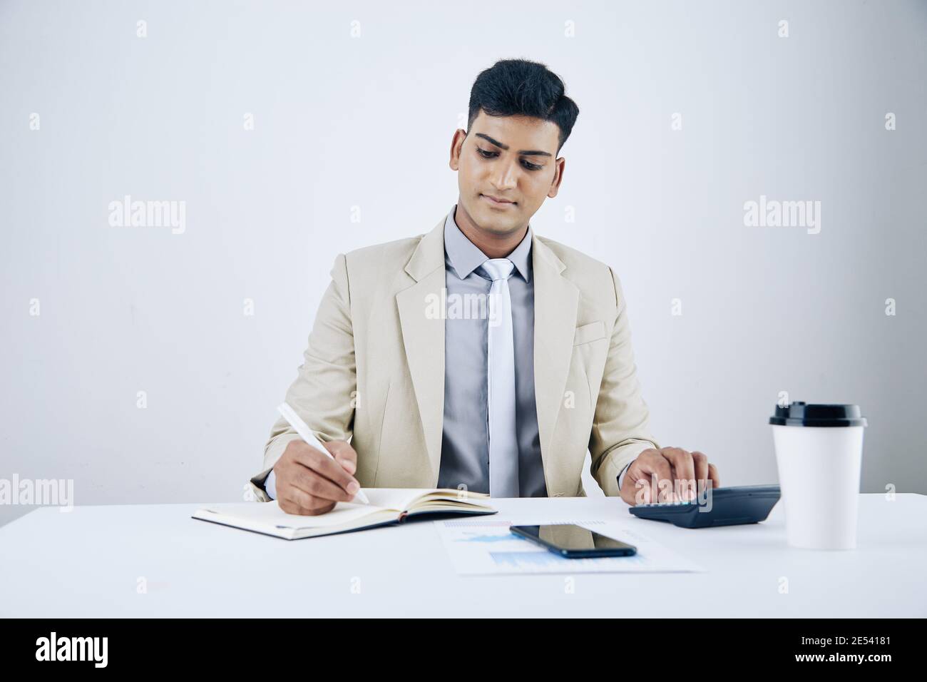Souriant, beau jeune homme d'affaires assis au bureau, faisant des calculs et écrivant dans le planificateur Banque D'Images