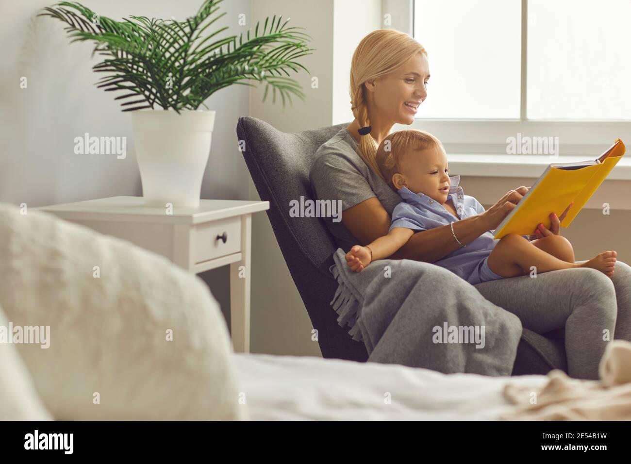 Bonne jeune maman lisant les premiers livres à son petit fils fauteuil dans une chambre confortable Banque D'Images