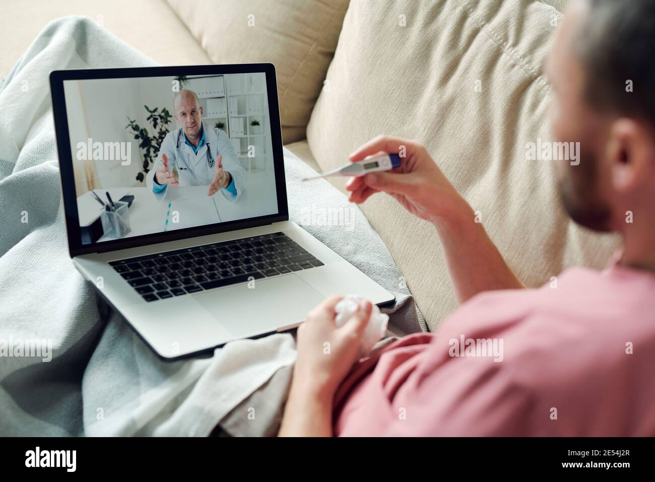 Homme malade avec ordinateur portable et mouchoir froissé assis sur un canapé sous la couverture tout en regardant le thermomètre et en consultant le médecin en ligne Banque D'Images