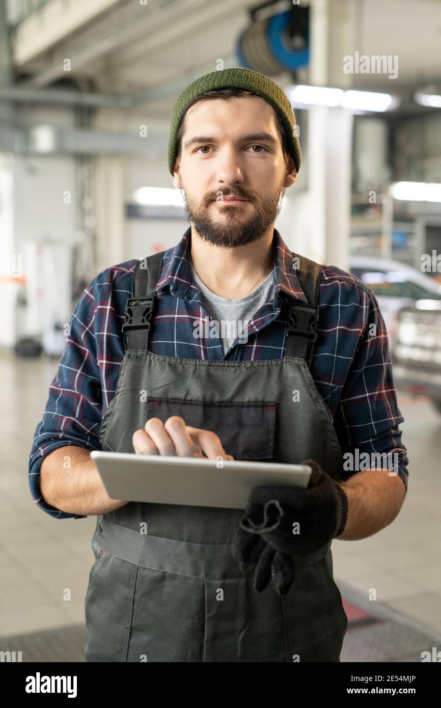 Jeune homme sérieux de service de voiture utilisant la tablette numérique tout en répondant aux questions en ligne des clients dans l'environnement de travail Banque D'Images