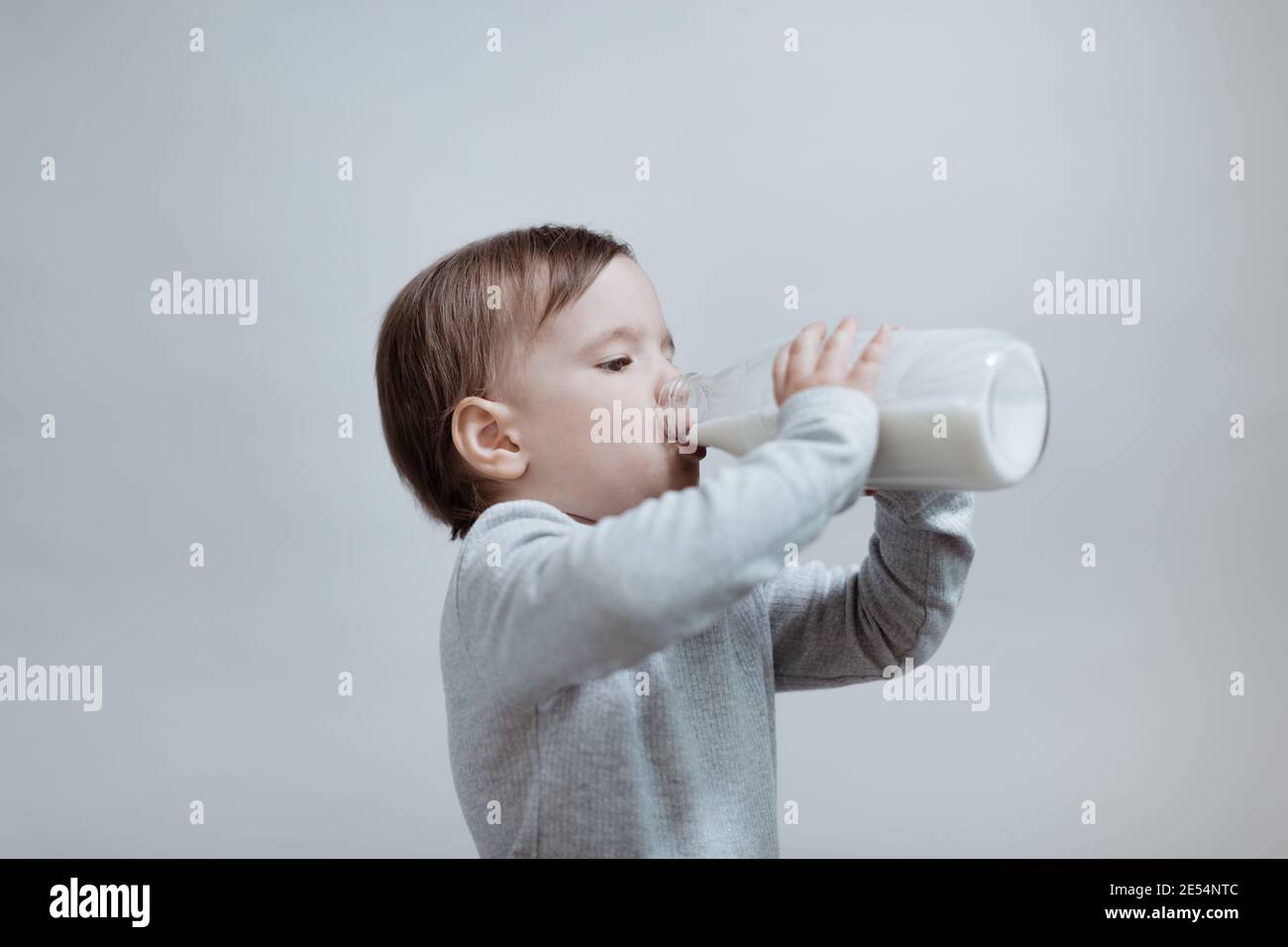 Un petit garçon boit du lait sur fond gris. Un portrait d'un jeune enfant tenant une bouteille de lait en verre. Banque D'Images