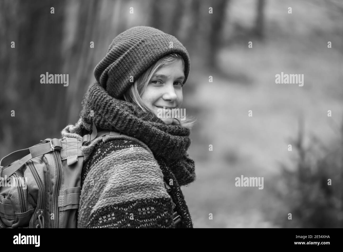 Portrait noir et blanc d'une jeune femme avec des couches de vêtements d'hiver lourds, un foulard en laine et un chapeau regardant l'appareil photo et souriant pendant une randonnée à la campagne. Banque D'Images