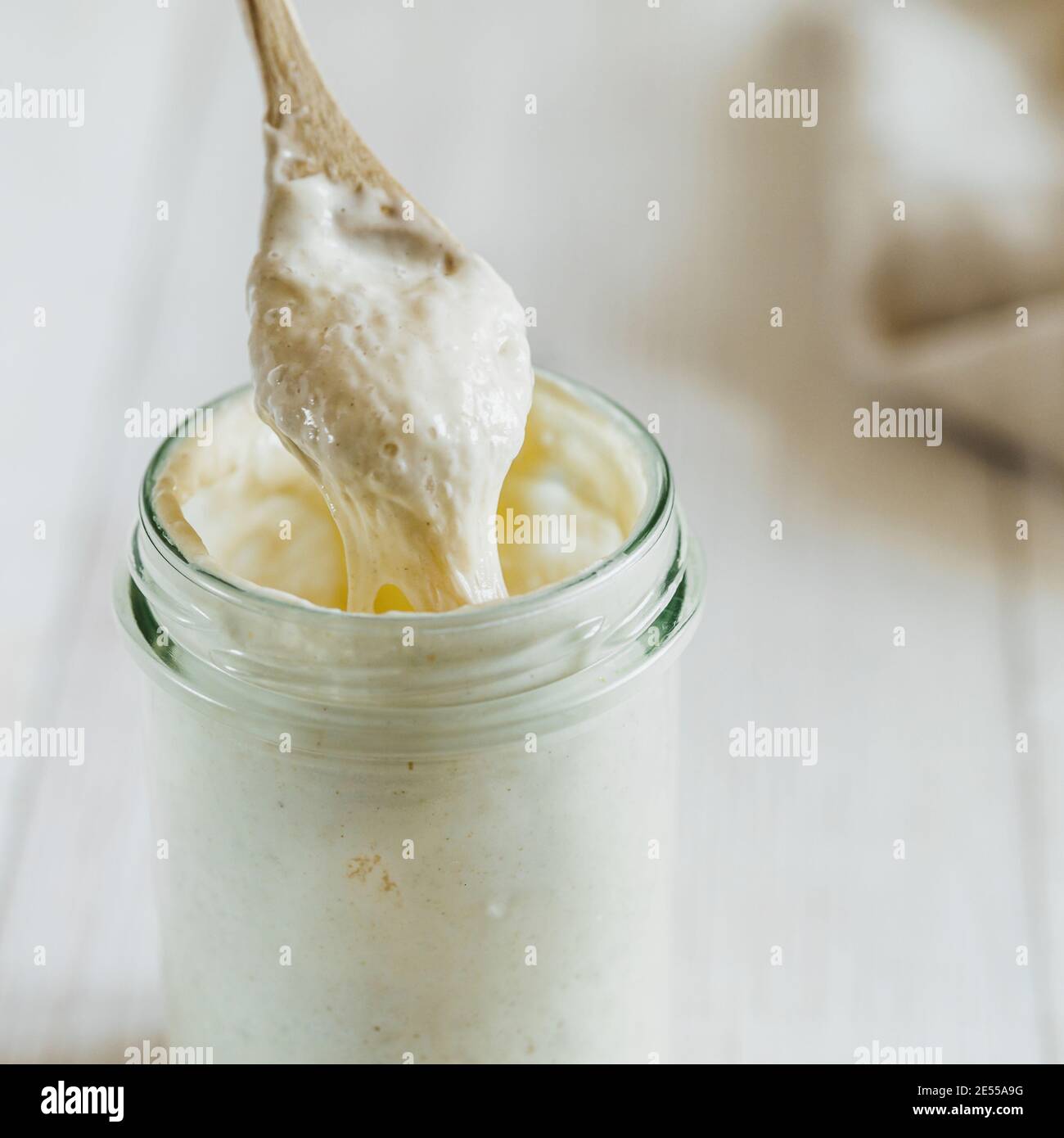 Prêt à préparer du pain de pâte de blé de démarrage goutte à goutte à partir d'une cuillère en bois. Pot en verre avec levain de départ 100 pour cent d'hydratation sur fond de bois blanc. Copier l'espace. Concept de pain maison Banque D'Images