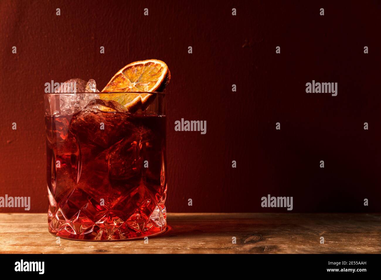 Verre avec cocktail glacé de type americano ou negroni avec entier tranche orange sur fond de mur rouge et rayon de lumière Banque D'Images