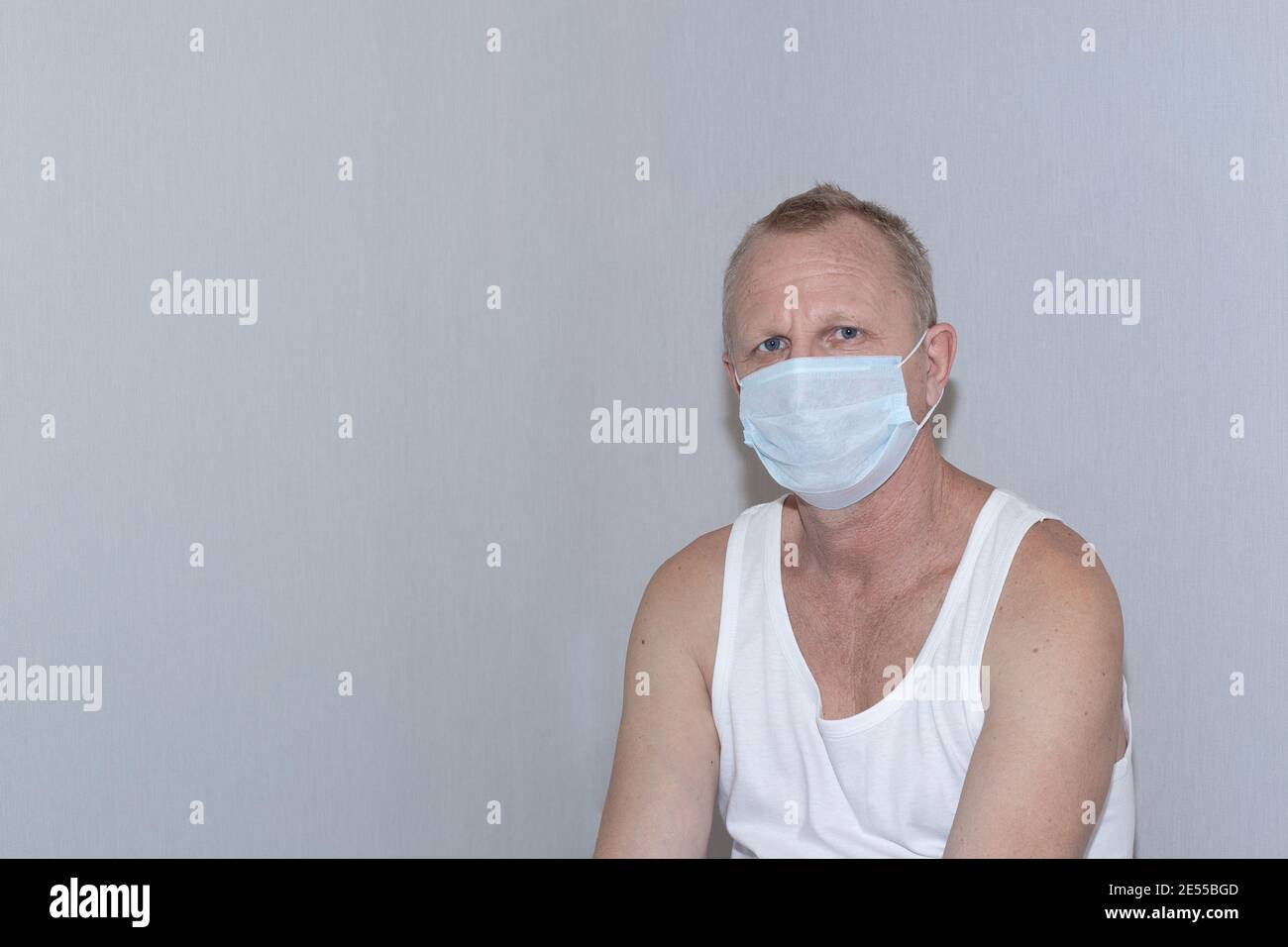 Homme d'âge mûr dans un T-shirt blanc et un masque de protection contre le fond d'un mur gris. Patient à l'hôpital. Banque D'Images