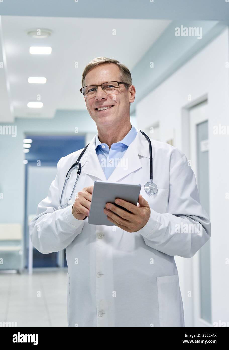 Un vieux médecin tenant une tablette numérique regardant la caméra à l'hôpital. Banque D'Images