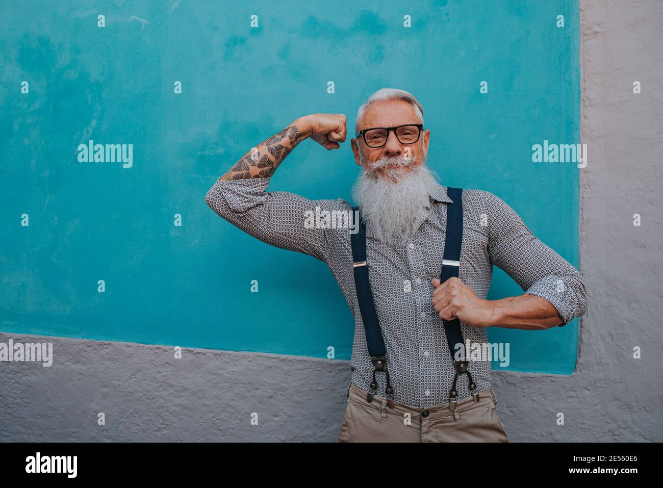 un homme plus âgé en vêtements et lunettes de taille basse et un la longue barbe blanche pose sur un mur bleu Banque D'Images