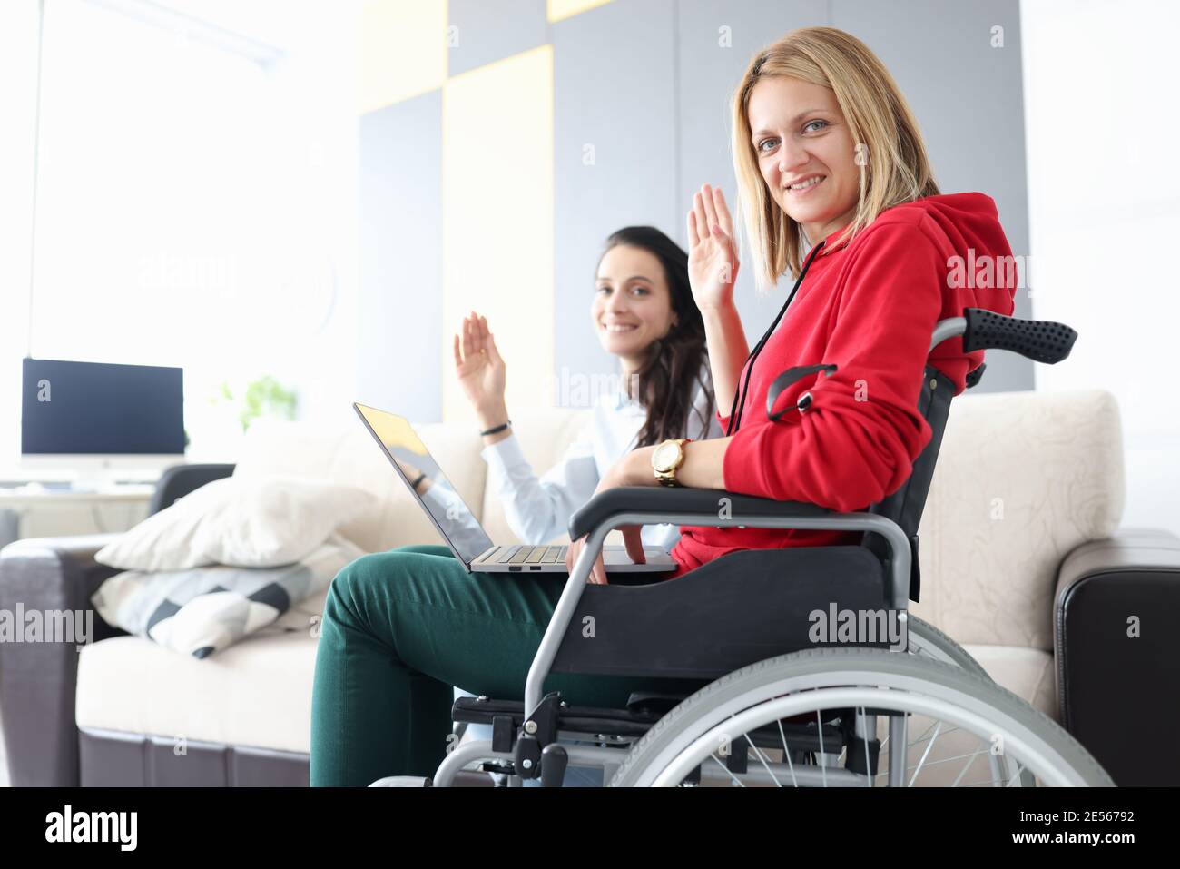 Femme en fauteuil roulant avec un ordinateur portable sur ses genoux agitant en saluant avec sa petite amie. Travail à distance pour les personnes handicapées concept de santé Banque D'Images