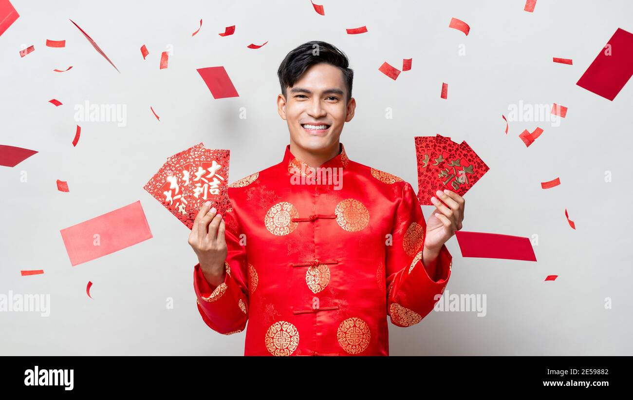 Souriant homme asiatique heureux en costume oriental traditionnel tenant des enveloppes rouges ou Ang Pao en fond gris avec confetti, le texte chinois signifie grand l Banque D'Images