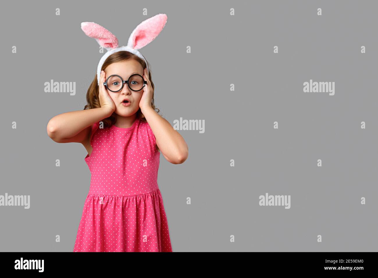Petite fille surprise portant des oreilles et des lunettes de lapin de Pâques. Portrait d'un enfant sur fond gris. Banque D'Images