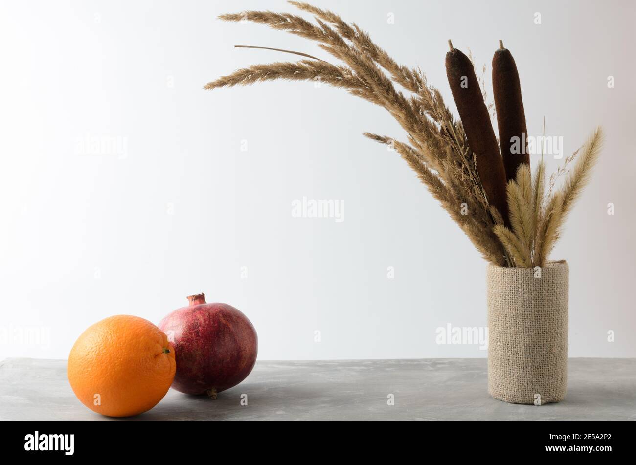 Une orange et une grenade sur une table grise texturée contre un mur blanc avec un bouquet d'automne d'herbes séchées et de roseaux. Mise au point sélective. Banque D'Images
