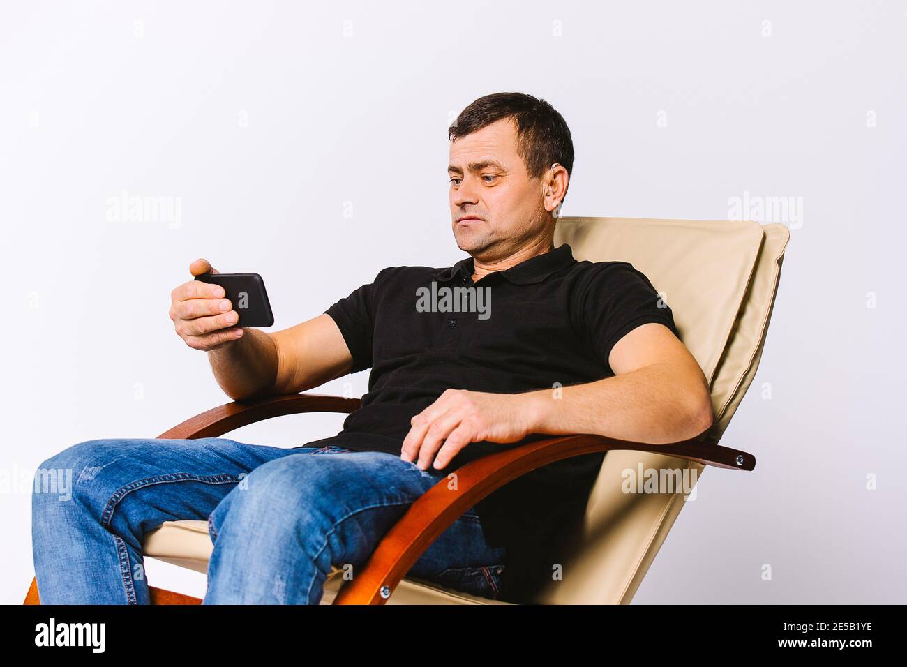 Homme sourd et âgé avec une prothèse auditive assis dans un fauteuil en cuir et regardant un film au téléphone. Arrière-plan blanc. Banque D'Images