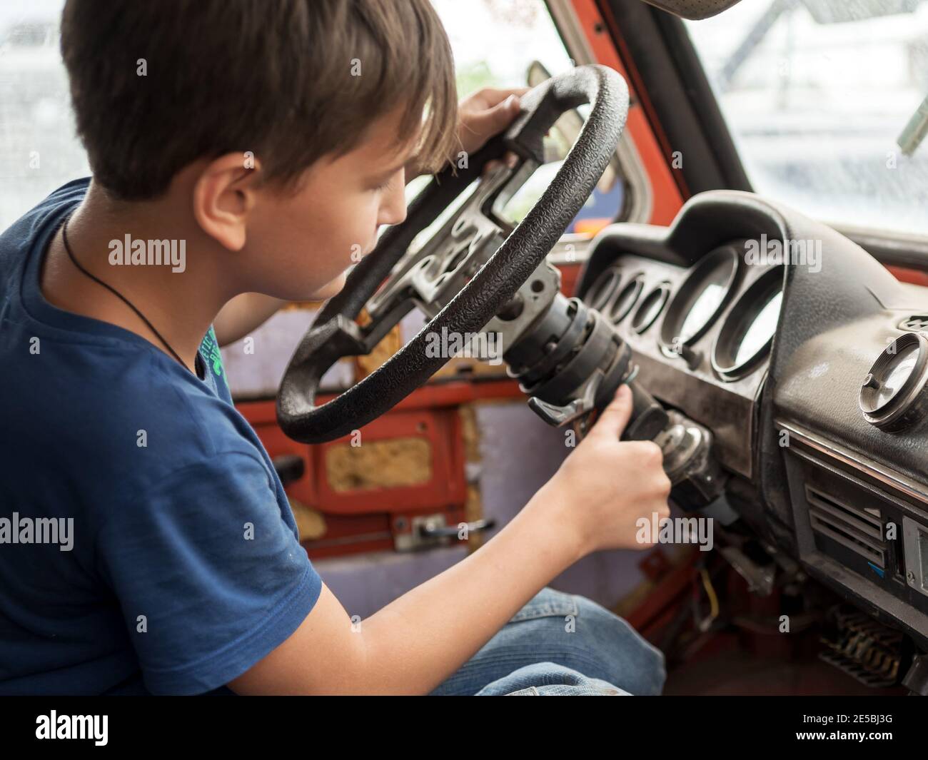 Un garçon de 11 ans joue avec enthousiasme le conducteur derrière le volant d'une vieille voiture abandonnée. Banque D'Images