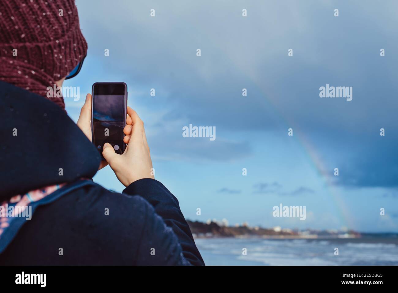 Retour Voir un homme dans des vêtements chauds prendre un téléphone photo de la vue de l'arc-en-ciel au-dessus de la mer pendant une promenade sur la mer d'hiver. Unité avec la nature, plaisirs simples Banque D'Images