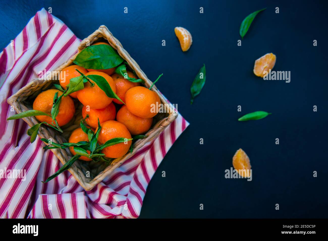 Vue du haut mandarines fraîches mûres, clémentine, mandarine avec feuilles vertes dans le bol de paille sur une serviette rayée et fond de pierre bleu foncé. Local Banque D'Images